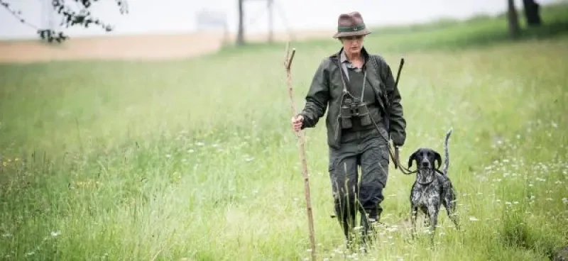 Sie sehen ein Bid Mit einem Jäger in passender Kleidung und einem Jagdhunf über ein Feld wandern. – Foto Foto: © Kultur_und_Tradition_Jagdfakten_Osterreich_Sliderbild_1960x900-600x276.jpg